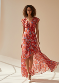 Woman wearing a red floral dress standing in a sunlit room with a neutral wall.